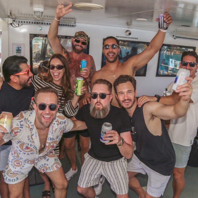 A group of friends on a boat raise drinks and smile for the camera, wearing sunglasses and casual summer clothes, enjoying a lively and sunny day by the water.