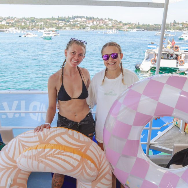 Two smiling women stand on a boat holding large inflatable pool rings, with one wearing a black bikini and the other in a white shirt and sunglasses; the sea, boats, and distant shoreline are visible in the background.