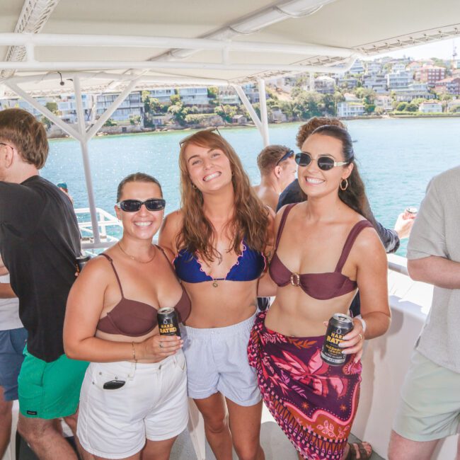 Three women wearing swimsuits and sunglasses smile and hold canned drinks on a boat, with sunny water and buildings visible in the background. Other people are socializing around them.