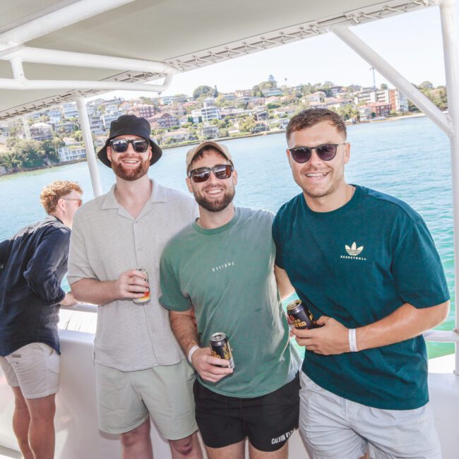Three men in casual summer clothing and sunglasses smile while holding drinks on a boat. The background shows water and buildings onshore under a clear sky. Other people are also present, enjoying the sunny day.