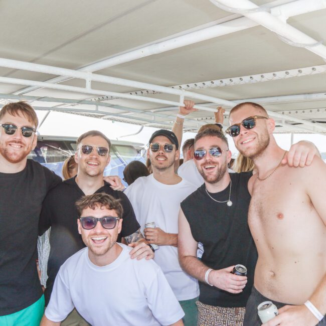 A group of six young men wearing sunglasses pose and smile for a photo on a boat. Some hold drinks, and they're surrounded by other people, with water and boats visible in the background.