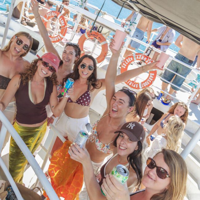 A group of smiling women hold drinks and pose for a photo on a sunny boat party, with people dancing and relaxing in swimwear in the background, surrounded by blue water and life rings.