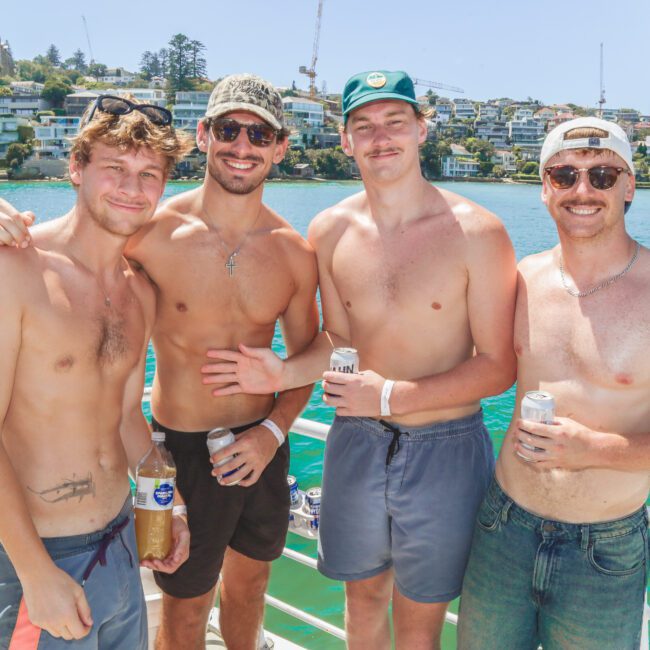 Four young men in swim trunks and sunglasses stand shirtless on a boat, smiling and holding drinks. Behind them is a scenic waterfront with houses, trees, and a sunny blue sky.