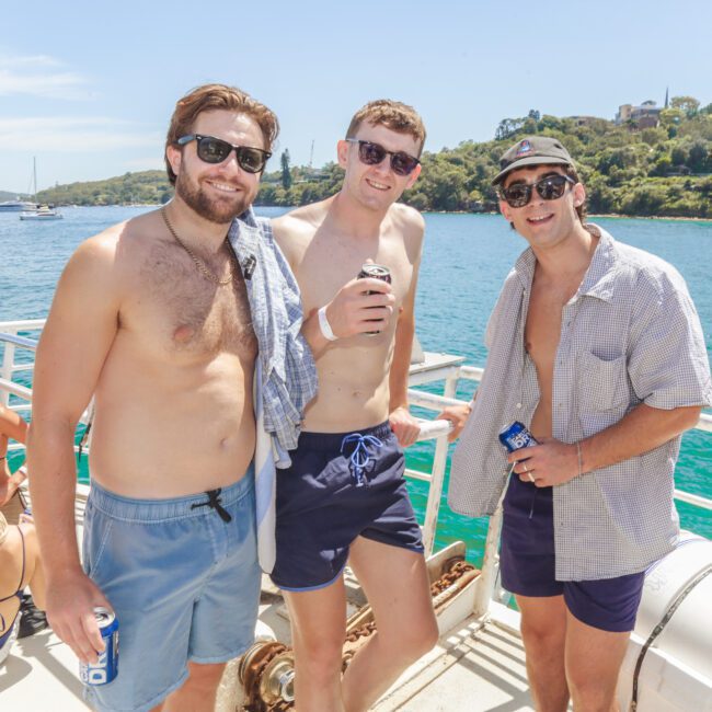 Three men wearing sunglasses and swim trunks pose together on a boat, each holding a drink. Sunny weather, clear blue water, and a scenic shoreline with houses are visible in the background. Other people relax nearby.