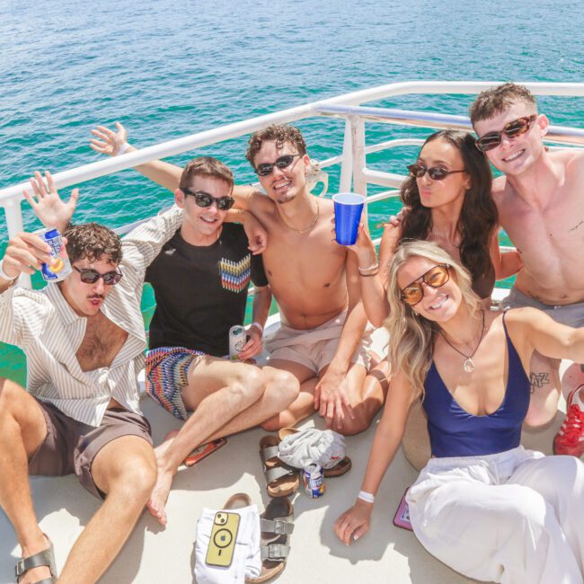 Six young adults sit on a boat deck, smiling and posing with drinks in hand. The group is casually dressed in swimwear and sunglasses, with the ocean and blue sky visible in the background.