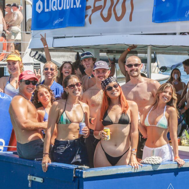 A group of people in swimsuits smile and pose together on a boat during a sunny day, holding drinks. A "LIQUID I.V." and "Yacht Social Club" banner is visible in the background.