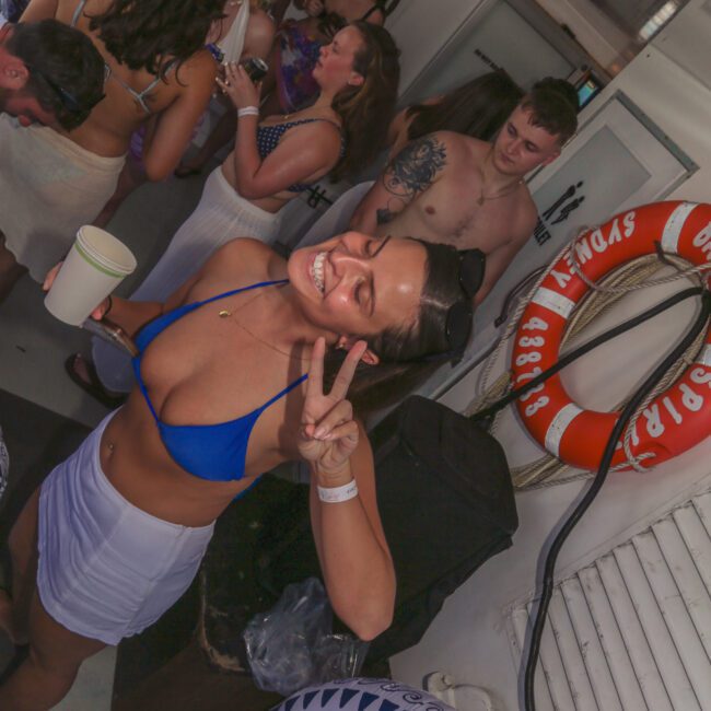 A woman in a blue bikini top and white skirt smiles and makes a peace sign at a lively boat party with other people in swimwear. Inflatable swim rings and life preservers are visible in the background.