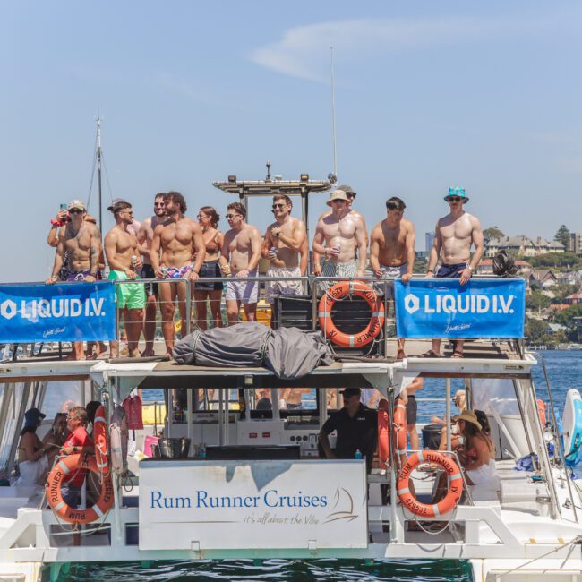 A group of shirtless men in swim trunks stand on the upper deck of a Rum Runner Cruises boat on the water, with city buildings and other boats in the background. Blue LIQUID I.V. banners hang on the boat.
