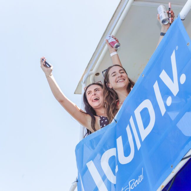 Two smiling women hold drinks and raise their arms while standing on a balcony decorated with a blue “LIQUID I.V.” banner, enjoying a sunny day at what appears to be an outdoor event.