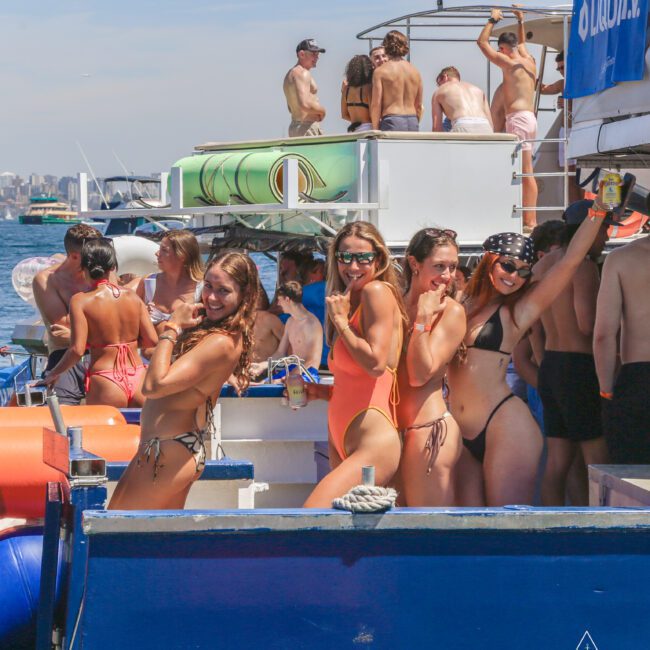A group of people in swimsuits enjoys a sunny day on a crowded boat. Several women in the foreground smile and pose for the camera, while others are socializing in the background near the water.