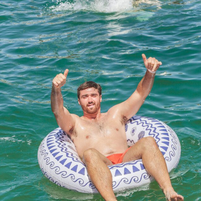 A man in orange swim trunks relaxes on a patterned inflatable tube in clear blue water, giving double thumbs-up. Other people swim and float in the background. "Yacht Social Club" logo is visible on the bottom right.