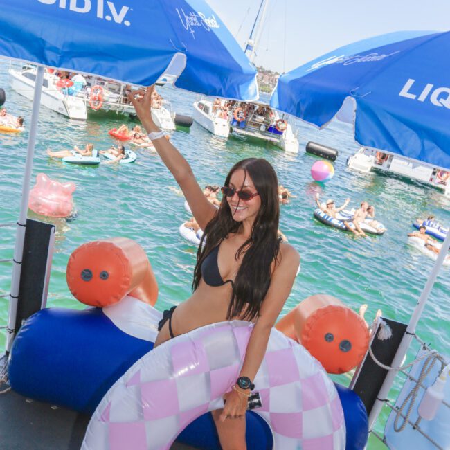 A woman in a black bikini and sunglasses smiles and poses with a pink checkered pool float under blue “Liquid I.V.” umbrellas on a dock, with boats and people swimming in the water behind her.