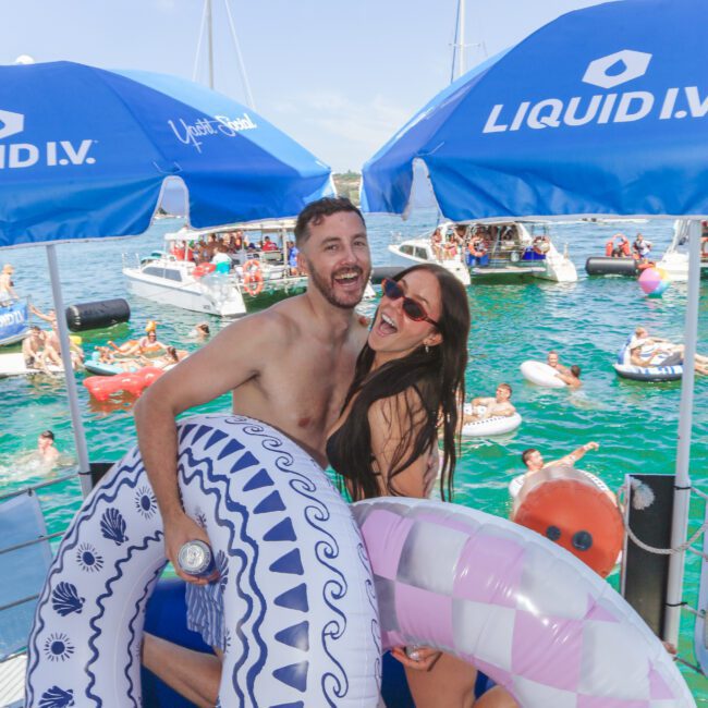 A smiling man and woman, both in swimsuits, pose with inflatable pool rings under blue umbrellas on a dock. Behind them, people relax on boats, inflatables, and in the water at a lively party.