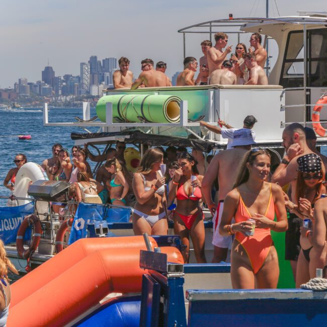A group of people in swimsuits enjoy a lively party on boats docked together on the water, with a city skyline visible in the background under a sunny sky.