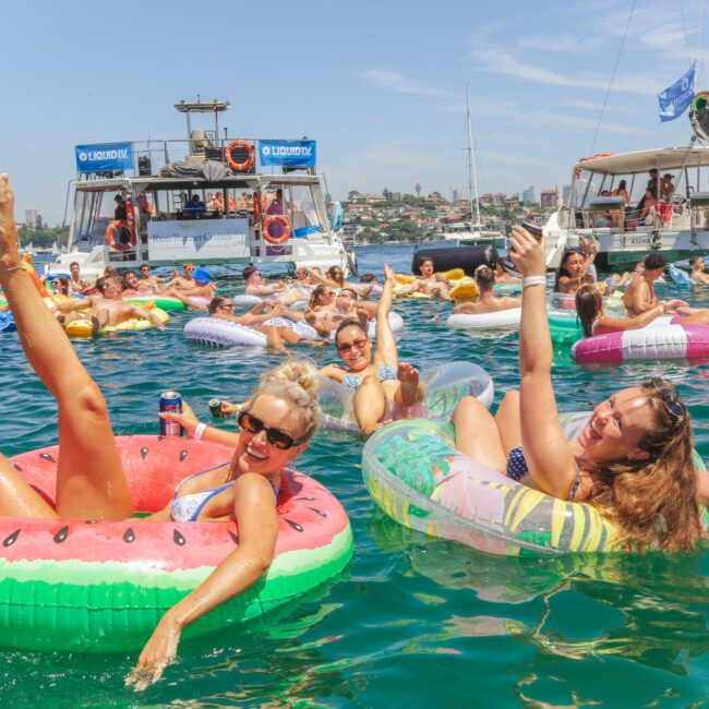 People relaxing on colorful pool floats in the ocean, smiling and raising drinks, with boats and more people in the background under a sunny blue sky. The atmosphere is festive and lively.