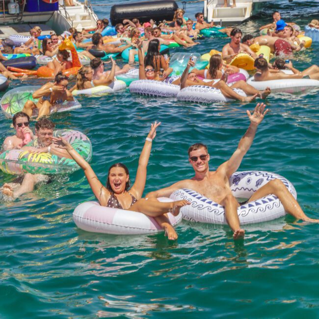 Dozens of people relax on colorful pool floats in a bright blue sea near several boats, smiling and raising their arms, enjoying a lively summer party under sunny skies.