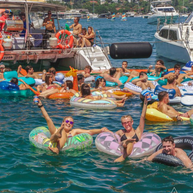 A lively group of people float on colorful inflatables in a blue harbor near anchored boats, enjoying the sunny weather with smiles and drinks. Green hills and houses are visible in the background.