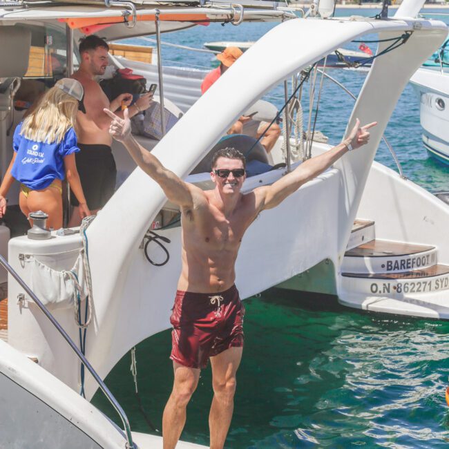 A man in red swim trunks stands at the back of a boat with arms raised and smiling. Other people are visible on the boat, and water and houses are in the background. It's a sunny day.