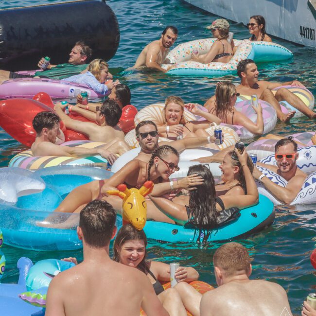 A group of people in swimsuits relax on colorful inflatable pool floats in the water near a boat on a sunny day, laughing and socializing.
