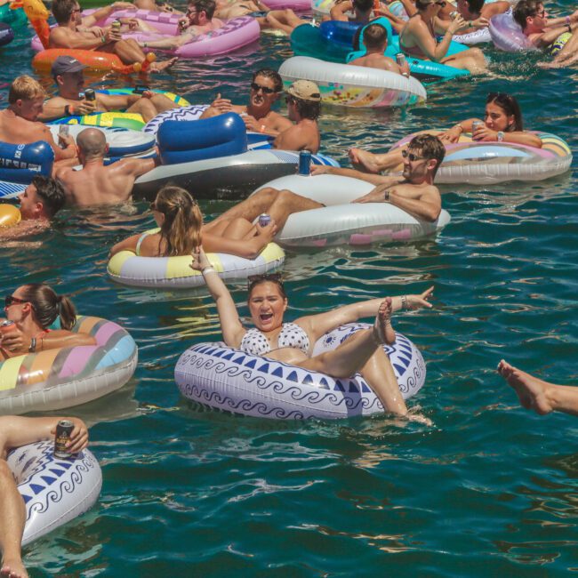 A large group of people relax on colorful inflatable pool floats in a crowded, sunlit body of water, laughing, talking, and enjoying drinks during a lively summer party.
