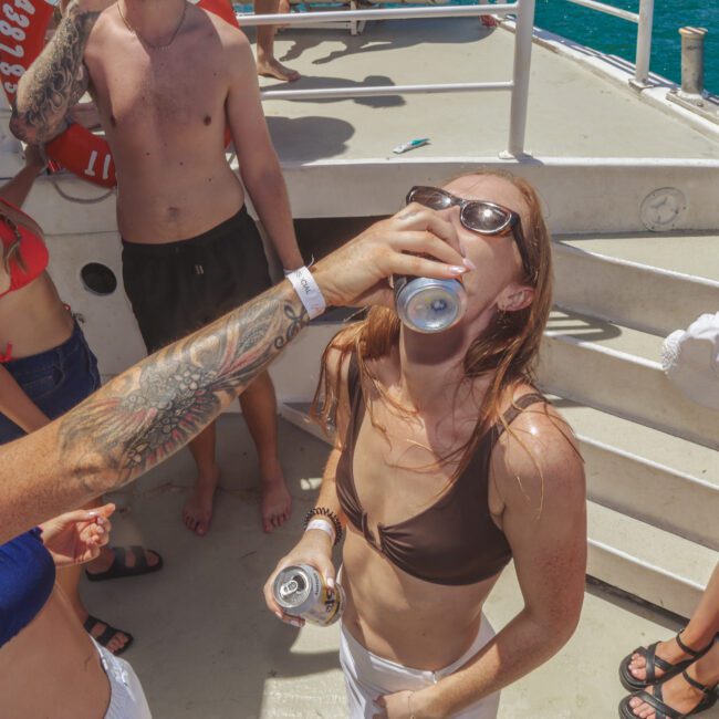 A woman in a brown bikini drinks from a can held by another woman with a tattooed arm on a boat. Several people in swimwear are laughing and enjoying the sunny day near blue water.