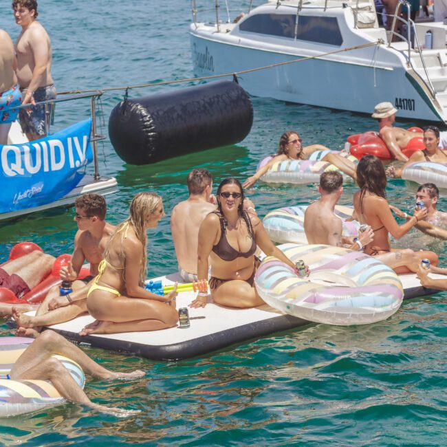 A group of young adults in swimsuits relax and socialize on inflatable floats and a floating dock in clear turquoise water near a boat, enjoying a sunny day. A banner with "LIQUIDIV" is displayed nearby.