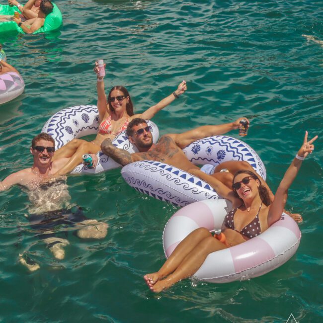 Four people float and relax on inflatable rings in clear blue-green water, smiling and making peace signs. Others float in the background. Sunlight glistens on the water, creating a lively, fun atmosphere. Logo reads “Yacht Social Club.”.