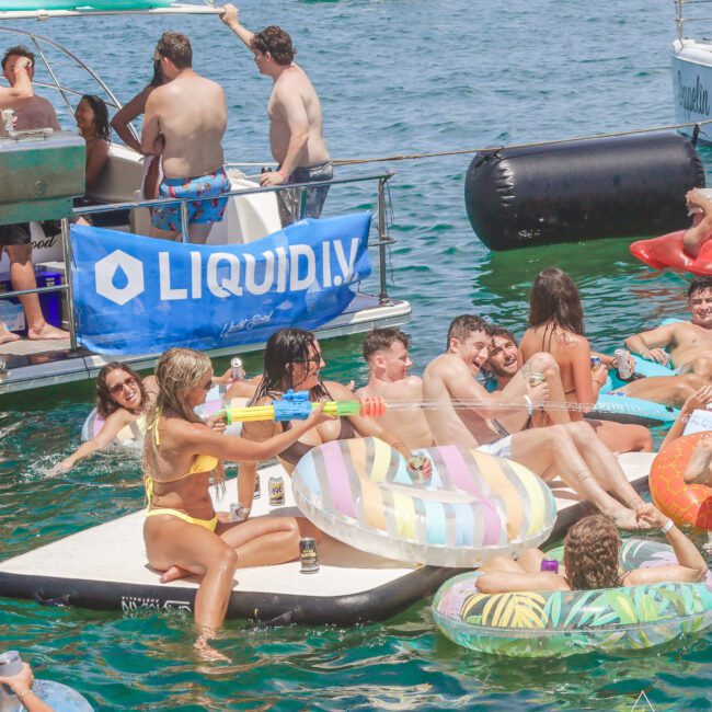 A group of people relax on colorful inflatables and paddleboards in a lake near a boat with a “Liquid IV” banner, enjoying sunny weather, drinks, and a festive summer atmosphere.