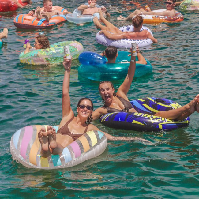 Dozens of people float in colorful pool tubes on sunny water; two women in front, smiling, hold drinks and wave, enjoying the lively summer atmosphere. The “Groove Social” logo appears in the corner.