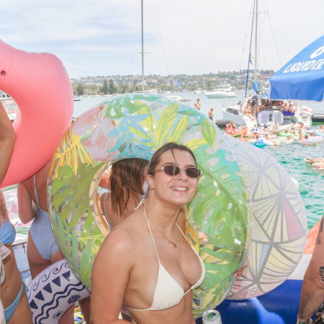 People in swimsuits smile and relax on a dock with colorful inflatable rings. Many others float in the water nearby. Boats and umbrellas are visible in the background on a sunny day.