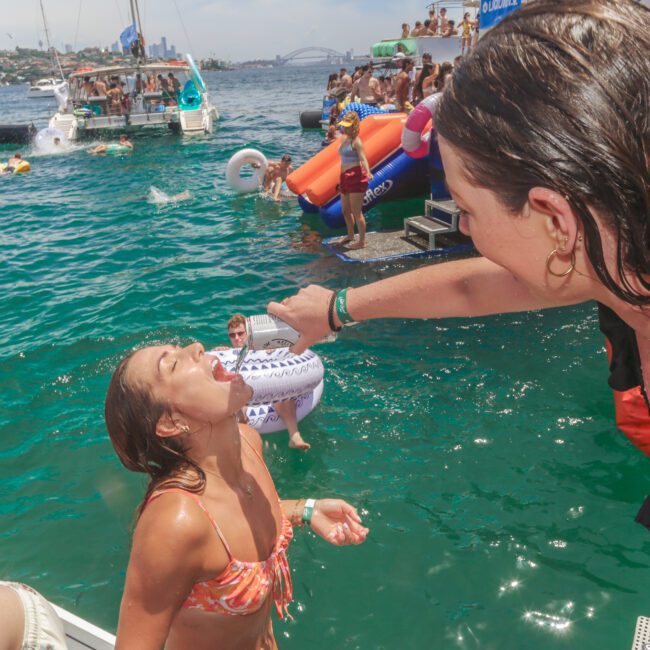 Two women at a lively boat party; one is pouring a drink into the other's mouth as they laugh beside the water, with people swimming, inflatables, and boats visible in the background under a sunny sky.