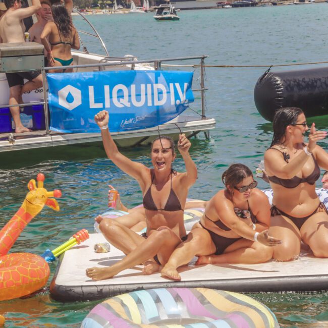 A group of people in swimsuits relax and cheer on a floating platform in a lake, surrounded by inflatable toys. A boat with a “Liquid IV” banner is nearby, and others swim and enjoy the sunny day in the background.