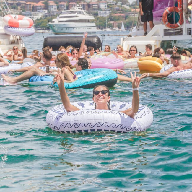 People relax on colorful inflatable rings in the water near boats on a sunny day. A smiling person in sunglasses and a white floatie flashes a peace sign. Crowds and yachts are visible in the background.