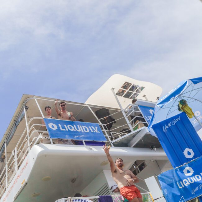 Three people enjoy a sunny day on a boat decorated with blue “LIQUID I.V.” banners; two stand on the upper deck waving, while one in orange shorts greets from below near a blue umbrella. The sky is clear and bright.