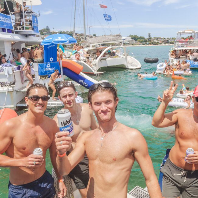 Four smiling men in swimwear hold drinks and pose for a photo on a boat, with several other people and boats in the background enjoying a sunny day on the water. The atmosphere is festive and lively.