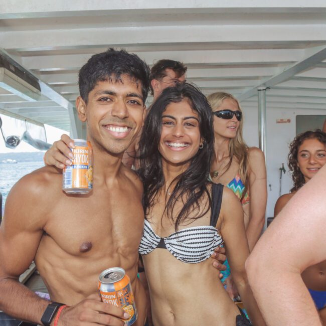 A group of young adults in swimsuits smile and pose together on a boat; two in front hold canned drinks, and the sea is visible in the background.