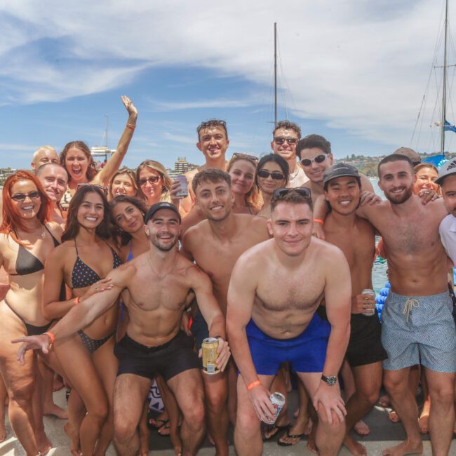 A large group of people in swimwear smile and pose together on a boat under a sunny sky, with yachts and blue water in the background. Some hold drinks, and everyone appears to be enjoying a fun day outdoors.