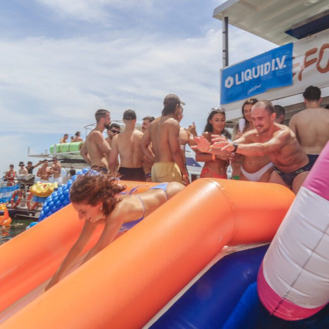 A group of people in swimsuits enjoy a party on a boat; one woman slides down an inflatable slide into the water while others watch and cheer. The scene is lively and festive under a sunny sky.