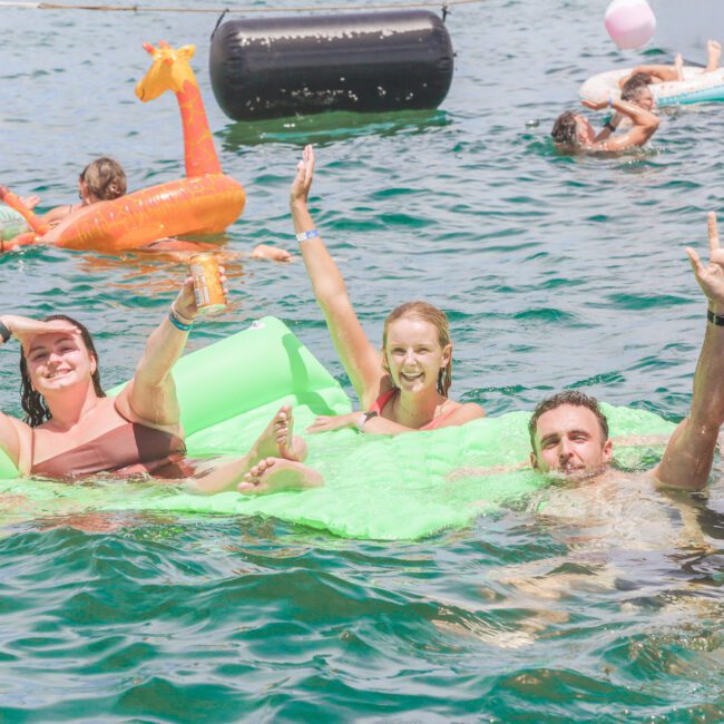 Five adults smile and wave while floating together in clear blue water, some holding onto a bright green inflatable. Other people on colorful floaties can be seen in the background, enjoying a sunny day on the water.