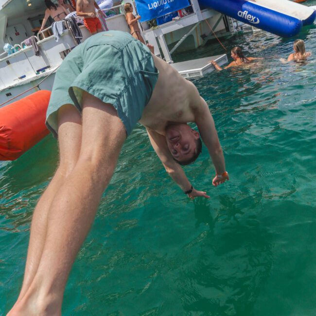 A man in green swim shorts dives into clear blue water from a boat, with other people swimming and relaxing on the boat in the background. Banners and inflatables are visible, creating a lively summer atmosphere.