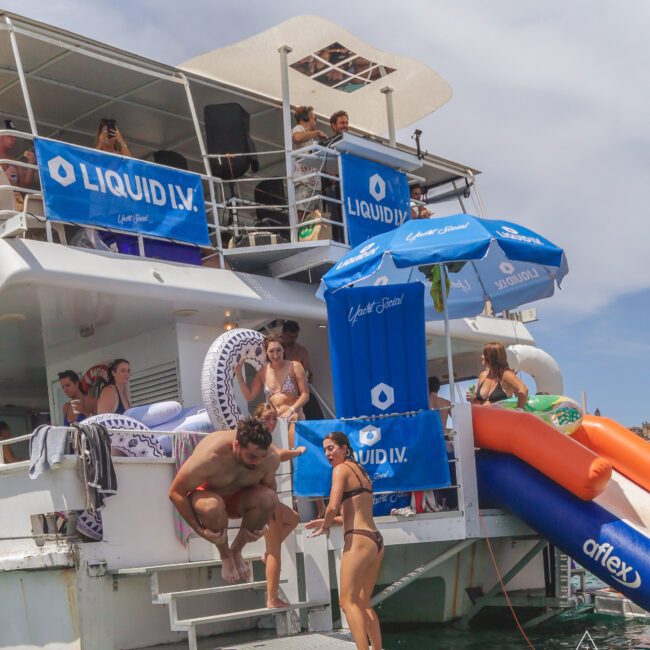 A group of people enjoy a sunny day on a yacht, with some standing on the boat and others using a slide into the water. Blue banners and umbrellas with “LIQUID I.V.” branding are visible on the yacht.