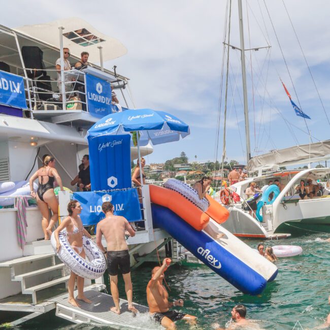 People enjoy a lively yacht party, swimming, holding pool floats, and using inflatable slides off the boat into the water. The scene is sunny, with other boats and a coastal backdrop visible.