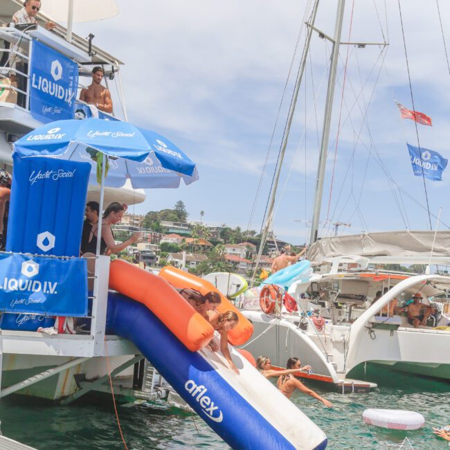 People slide down a dual inflatable slide from a boat into the water at a lively boat party. Others watch from the deck. Blue Liquid I.V. banners and branded decor are visible. Another boat and houses appear in the background.