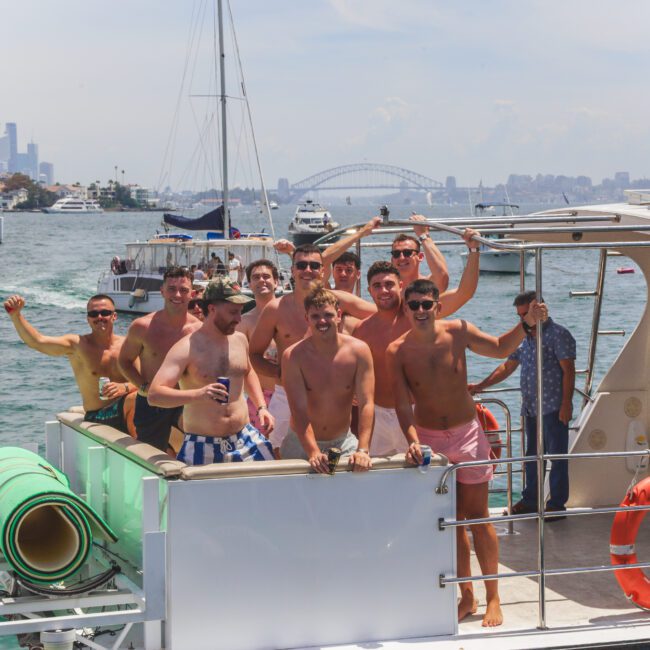 A group of smiling men in swim trunks pose on the deck of a boat on a sunny day, with a city skyline, bridge, and other boats visible in the background.