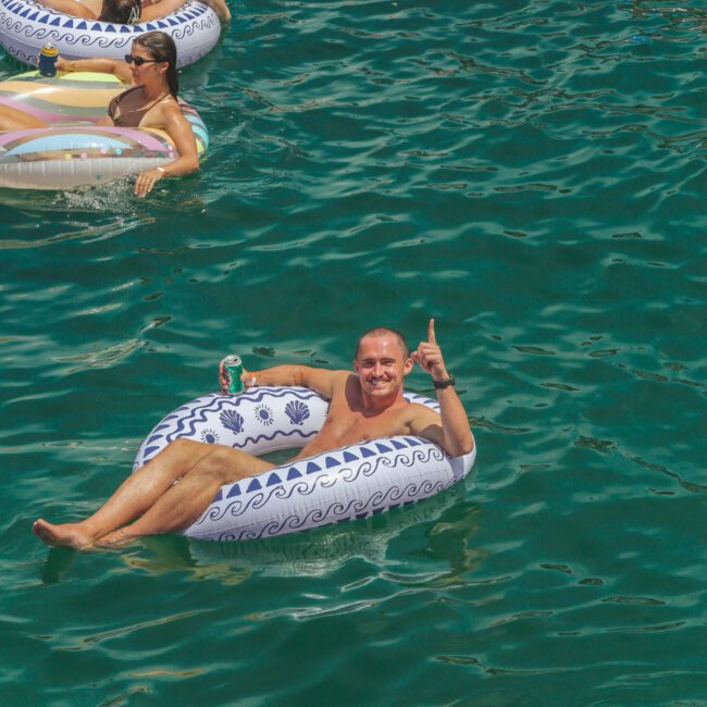 A group of people float on inflatable tubes in clear blue water. One man in front smiles and gives a thumbs-up to the camera while holding a drink. The sun is bright, and everyone appears relaxed and happy.