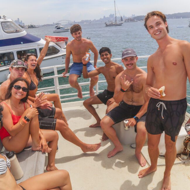 A group of young adults in swimwear smile and pose on a boat deck, holding drinks and snacks, with other boats and a city skyline visible in the background on a sunny day.