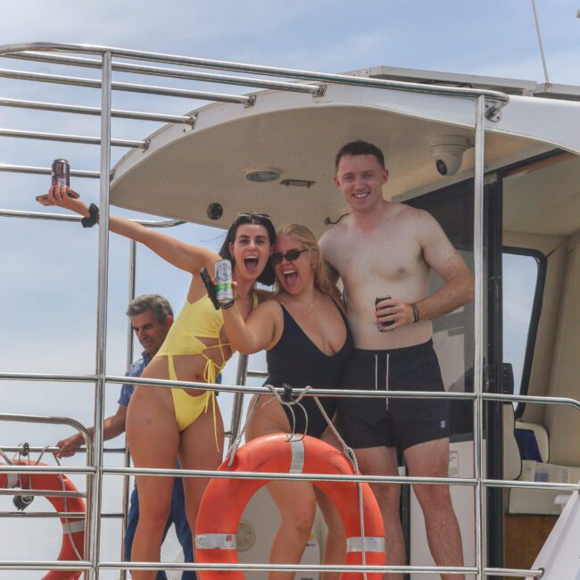 Three people in swimsuits pose and smile on the deck of a boat, holding drinks. Two orange life rings are visible, and a fourth person stands in the background. The sky is partly cloudy.