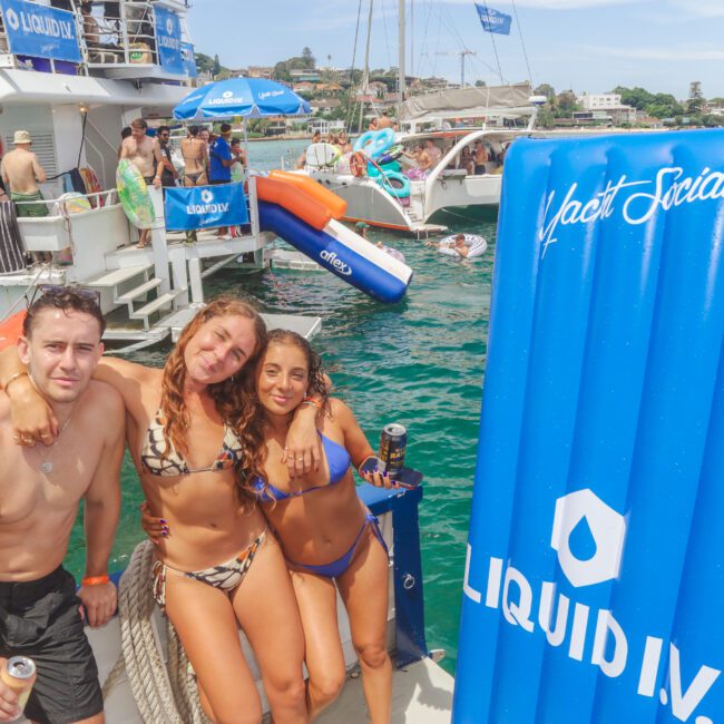 Three people in swimsuits smile and pose on a boat, with other partygoers, boats, and inflatable floats in the background. Branded “Liquid IV” and “Yacht Social” banners are visible. The weather is sunny and festive.