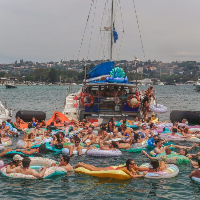 A large group of people relax on colorful inflatable pool floats in the water near several boats during a lively outdoor party. The background shows houses on a hillside under a cloudy sky.