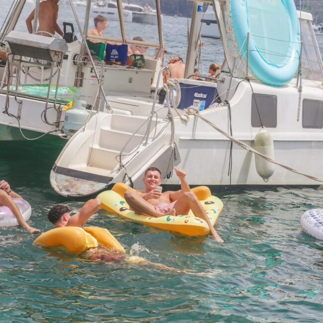 People relaxing on inflatables in the water next to a white yacht, with one man smiling and lounging on a yellow float. Others are near the boat, and the background shows water and distant shore.